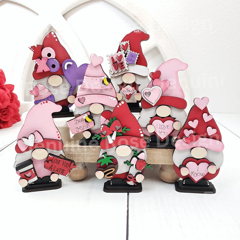 Puede incluir: Ocho gnomos de San Valent&iacute;n con diferentes dise&ntilde;os y lemas. Los gnomos est&aacute;n hechos de madera y pintados en tonos de rojo, rosa y blanco. Algunos de los lemas incluyen "Te amo", "S&eacute; m&iacute;o" y "Te amo un latte".