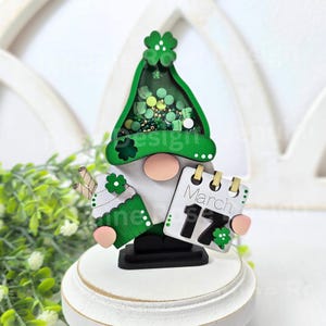 May include: A green and white gnome figurine holding a calendar with the date March 17th. The gnome's hat is filled with green, gold, and white glitter.