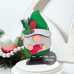 May include: A wooden Christmas gnome figurine with a green hat, white beard, and red mitten. The gnome has a round face and is decorated with holly and the word "HO HO HO". The base has the text "SVG Laser Cut File".