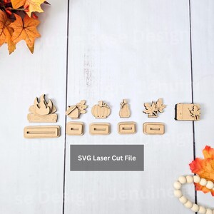 May include: A selection of laser-cut wooden autumn-themed shapes, including a campfire, acorn, pumpkin, pear, and maple leaves. Each shape has a matching base. The text "SVG Laser Cut File" is displayed on a grey rectangle.