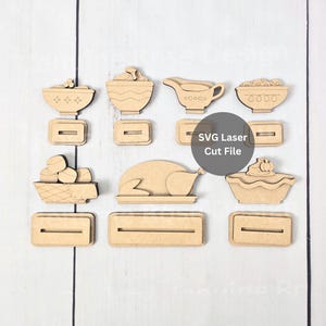 May include: A collection of laser-cut wooden shapes featuring Thanksgiving food items including a turkey, pie, gravy boat, and bowls, accompanied by rectangular stands with slots. The text SVG Laser Cut File is visible on the image.