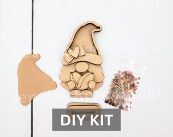 May include: A light brown wooden DIY kit featuring a gnome cutout with a heart detail, a separate hat piece, a rectangular base with a slot, and a small clear plastic bag containing assorted small brown and tan circular beads for decoration.