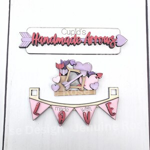 Op de afbeelding: Houten decoraties met Valentijnsdagthema. Bevat een bord met de tekst "Cupid's Handmade Arrows", een Cupido met pijl en boog en een banner met de tekst "made with LOVE". De decoraties zijn in roze, paarse en witte tinten.