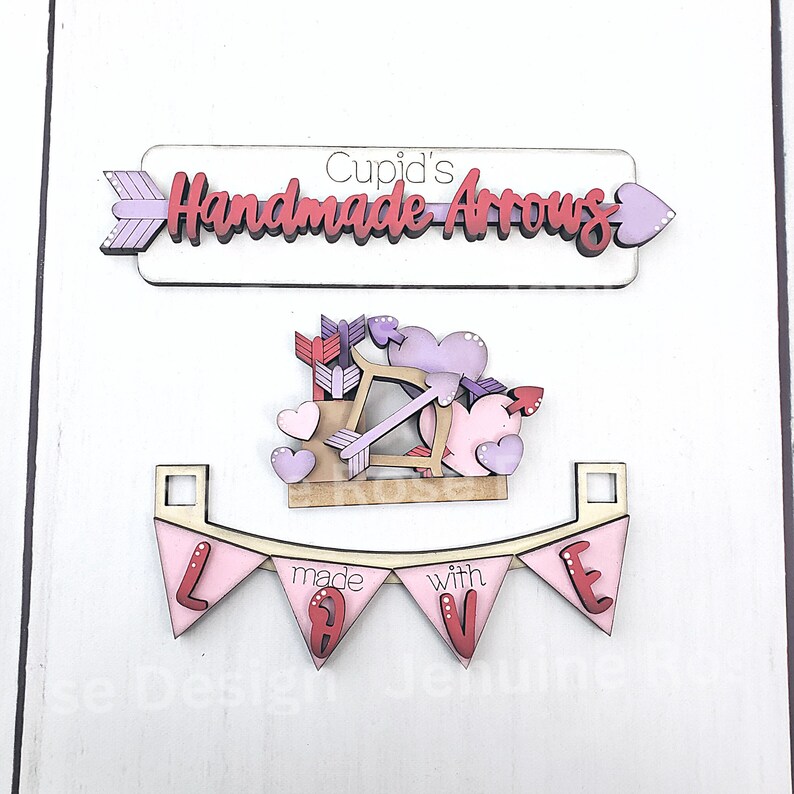 Op de afbeelding: Houten decoraties met Valentijnsdagthema. Bevat een bord met de tekst "Cupid's Handmade Arrows", een Cupido met pijl en boog en een banner met de tekst "made with LOVE". De decoraties zijn in roze, paarse en witte tinten.