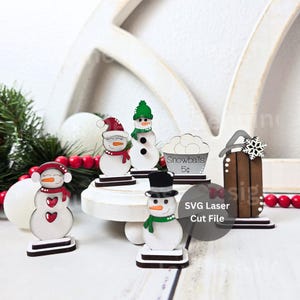 May include: Wooden snowman figurines with various accessories, including hats, scarves, and a bucket of snowballs. The figurines are on a white surface with a decorative wooden arch in the background. The text "Snowballs 5¢" is visible.