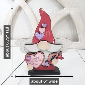May include: A wooden Valentine's Day gnome figurine. The gnome has a red hat, a white beard, and a pink face. It holds a heart-shaped sign that reads "Happy Valentine's Day." The figurine is about 6.75 inches tall and 6 inches wide.