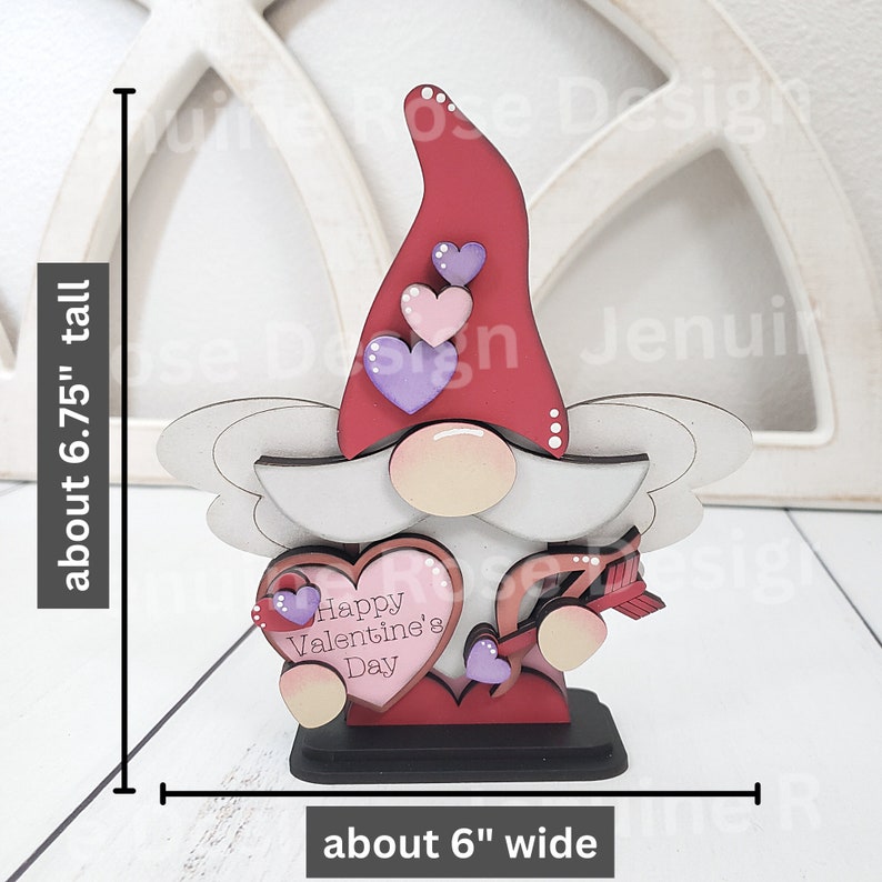 May include: A wooden Valentine's Day gnome figurine. The gnome has a red hat, a white beard, and a pink face. It holds a heart-shaped sign that reads "Happy Valentine's Day." The figurine is about 6.75 inches tall and 6 inches wide.