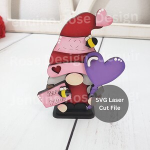 Puede incluir: Un gnomo de madera sosteniendo un globo de corazón morado y un cartel que dice "Bee Mine". El gnomo lleva un sombrero rojo con un sombrero a rayas rosa y blanco y una abeja en el sombrero. El gnomo está de pie sobre una base negra. Archivo de corte láser SVG.