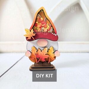 May include: A wooden gnome-shaped DIY kit with a fall theme. The gnome has a white face, a red and orange hat filled with decorative elements, and holds autumn leaves. The base has the text "DIY KIT".
