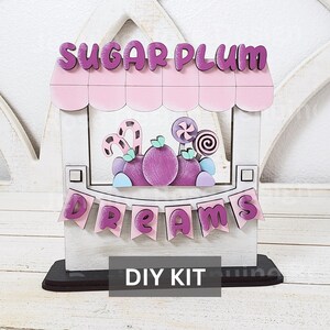May include: DIY kit for a wooden sign that says "Sugar Plum Dreams". The sign is painted white with pink accents and features a candy shop theme with purple and pink candy, lollipops, and a candy cane.