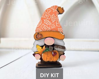 May include: A wooden gnome figurine with an orange hat patterned with leaves and coffee cups. The gnome holds a pumpkin with the word "Fall" and a coffee cup. The base of the gnome has the text "DIY KIT".
