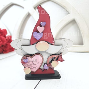 May include: A wooden Valentine's Day gnome figurine. The gnome has a red hat with three heart accents, a white beard, and a pink nose. It holds a heart-shaped sign that reads "Happy Valentine's Day". The figurine is on a black base.
