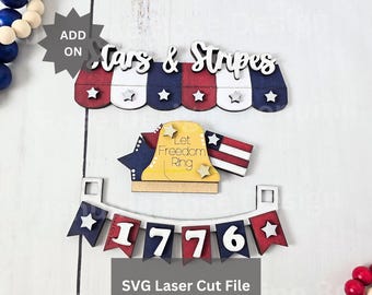 May include: A set of laser-cut wooden pieces featuring a red, white, and blue awning with the text Stars & Stripes, a yellow bell with the text Let Freedom Ring, and a banner with the text 1776, accompanied by small wooden stars and beads.