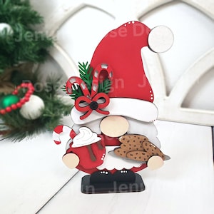 May include: A wooden Christmas gnome holding a red mug with whipped cream and a candy cane. The gnome is wearing a red hat with a white pom-pom and a white beard. The gnome is also holding a plate of chocolate chip cookies.