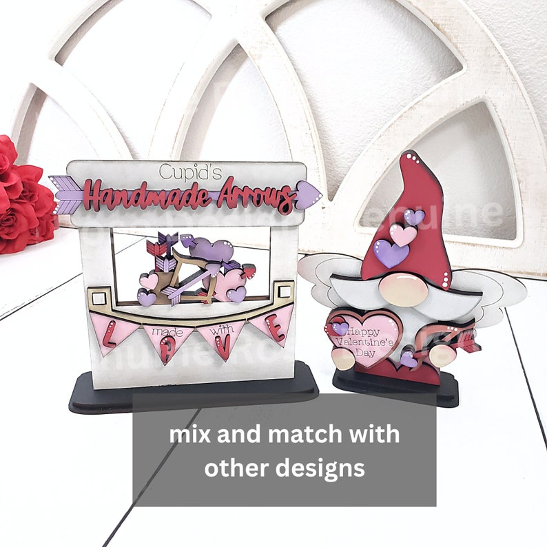 Op de afbeelding: Twee houten decoraties met Valentijnsthema. E&eacute;n is een bord met "Cupid's Handmade Arrows" met pijlen en harten. De andere is een kabouter met een hart met "Happy Valentine's Day".