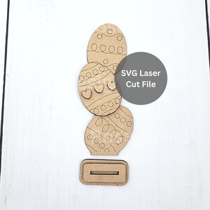 May include: A wooden Easter egg decoration set. The set includes four laser-cut eggs with decorative patterns and a base. The eggs feature etched designs of circles, hearts, and swirls. The text "SVG Laser Cut File" is visible.