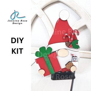 May include: DIY kit for a Christmas gnome craft project. The gnome is holding a red gift box with green wrapping paper and a candy cane. The gnome is wearing a red hat with white trim and a white beard. The gnome is also holding a glass of milk and a cookie.