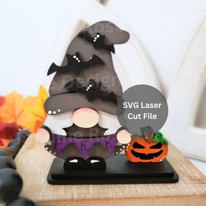 May include: A wooden Halloween gnome with a black hat with bats, a purple banner with the letters 'EKK', and a white beard. The gnome is holding a small orange pumpkin with a jack-o'-lantern face. SVG Laser Cut File.