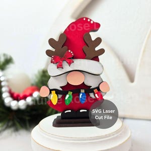 May include: A wooden Christmas gnome with a red hat, brown antlers, and a white beard. The gnome has a string of colourful lights and a red bow. The base is white and the text "SVG Laser Cut File" is on the side.