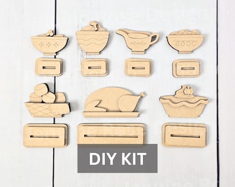 May include: A set of unfinished light brown wooden cutouts featuring Thanksgiving food items including a turkey, pie, and bowls, accompanied by matching rectangular slotted bases for display. The image includes a gray box with the text DIY KIT.