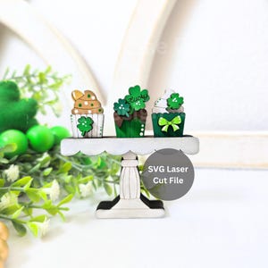May include: Three green and white cupcakes with shamrock and bow decorations on a white cake stand. The cupcakes are decorated with green and gold sprinkles. The stand has a scalloped edge. The text "Lucky" is on one of the cupcakes. SVG Laser Cut File.