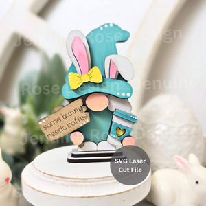 Könnte beinhalten: Ein türkisfarbener Zwerg mit einem rosa-weißen Hasenhut und einer gelben Schleife. Der Zwerg hält eine Kaffeetasse und ein Schild mit der Aufschrift "some bunny needs coffee".