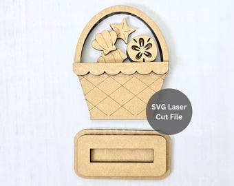 May include: A light brown laser-cut wooden basket shape featuring a handle and a cross-hatched pattern. Inside the basket are cutouts of a seashell, a starfish, and a sand dollar. Below the basket is a rectangular wooden base with a slot. Text reads SVG Laser Cut File.