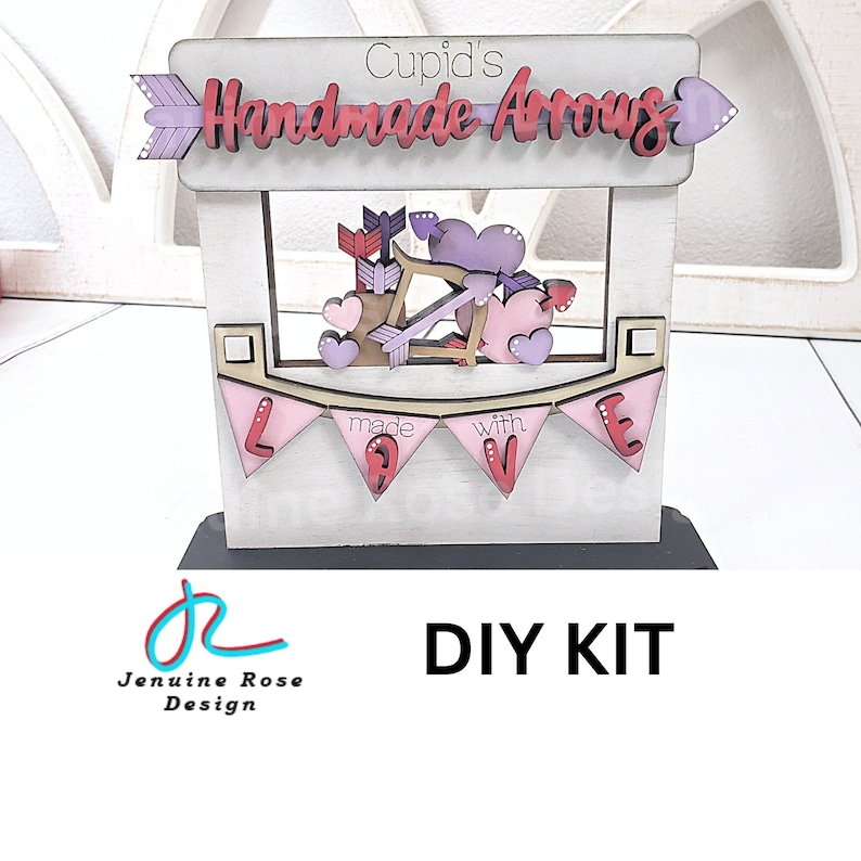 Op de afbeelding: Een houten doe-het-zelf kit voor Valentijnsdag. De display heeft een banner met de tekst "made with LOVE", een bord met "Cupid's Handmade Arrows" en decoratieve pijlen en harten in roze, rood en paars.