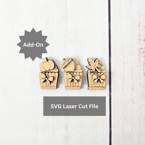 May include: Three laser-cut wooden cupcake-shaped designs with fall-themed toppers: a pumpkin, an acorn, and a maple leaf. Each cupcake has a bow detail. The text "Add-On" is above the cupcakes, and "SVG Laser Cut File" is below.