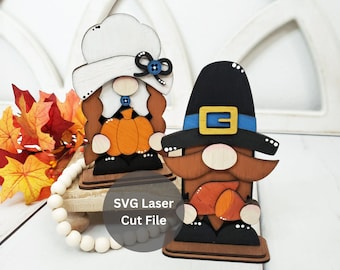 May include: Two wooden gnome figurines, one wearing a white hat and holding a pumpkin, the other wearing a black hat with a blue band and holding a corn cob. The gnomes are standing on wooden bases and the text "SVG Laser Cut File" is visible below.