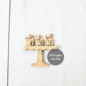 May include: Wooden Halloween cupcake cutouts on a decorative stand. Each cupcake is topped with a bat, skull, and spider, with the letters E, K, and K. The stand has a scalloped edge and a pedestal base. The text "SVG Laser Cut File" is also present.