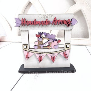 Op de afbeelding: Houten Valentijnsdagdecoratie met een bordje "Cupid's Handmade Arrows". De decoratie toont pijlen, harten en een pijl en boog. Een roze banner zegt "made with LOVE".