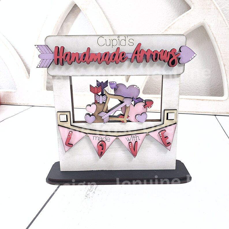 Op de afbeelding: Houten Valentijnsdagdecoratie met een bordje "Cupid's Handmade Arrows". De decoratie toont pijlen, harten en een pijl en boog. Een roze banner zegt "made with LOVE".