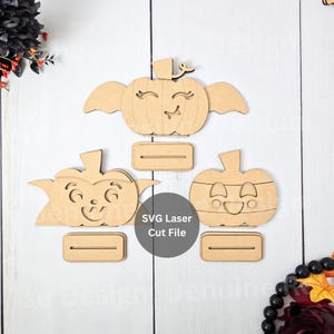Op de afbeelding: Drie houten pompoen uitsparingen met lachende gezichten en aparte bases. De pompoenen hebben verschillende ontwerpen, waaronder vleugels en verschillende gezichtsuitdrukkingen. De afbeelding bevat ook de tekst "SVG Laser Cut File".