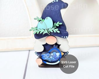 May include: A layered wooden gnome ornament featuring a dark blue hat with light blue dots, a peach-colored nose, white beard, and green foliage with two light blue eggs. The gnome holds a blue bird-shaped shaker filled with blue beads. Text reads SVG Laser Cut File.