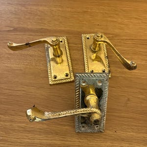 May include: Three antique brass door handles. Two are gold-toned, and one is silver-toned. Each handle has a rectangular backplate with a decorative rope-like border. The handles are curved and have a classic design.