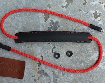 Könnte beinhalten: Roter Kameragurt mit einem schwarzen Lederpolster und zwei schwarzen Metallringen. Der Gurt besteht aus geflochtenem Seilmaterial. Das Lederpolster hat rote Nähte. Der Gurt liegt auf einer grauen Oberfläche mit einem braunen Lederanhänger, auf dem "Handmade Since 2005" steht.