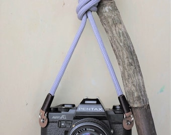 Könnte beinhalten: Ein graues Kameraband mit einer schwarzen Kamera daran. Die Kamera ist eine Pentax Super A mit einem 50mm Objektiv. Das Band hängt an einem Ast.