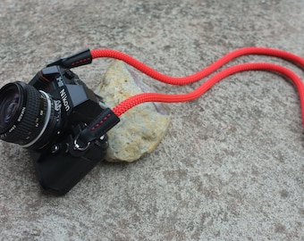 May include: A black Nikon camera with a red camera strap. The camera is sitting on a rock.