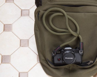 Könnte beinhalten: Eine schwarze Olympus-Kamera mit einem grünen und roten Kameragurt in einer grünen Stofftasche.
