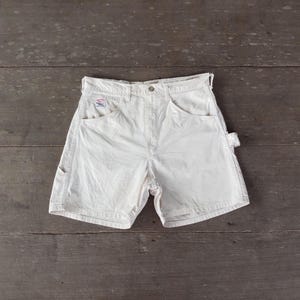 May include: Cream-colored work shorts with multiple pockets and a small logo on the front. The shorts have a classic design with a button and zipper closure. The shorts are made of a durable fabric, perfect for outdoor activities or casual wear.