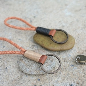 May include: Two leather keychains with braided straps and metal key rings. The keychains are on top of two gray stones. A small key with a metal chain is on the right side of the image.