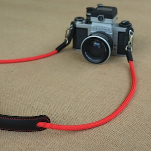 May include: A red camera strap with a black leather shoulder pad. The strap is made of a thick, durable material and has a simple, classic design.