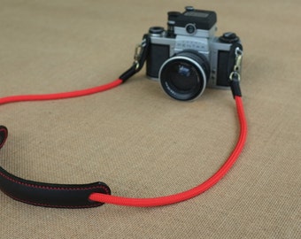Könnte beinhalten: Ein rotes Kameraband mit einem schwarzen Leder-Schulterpolster. Das Band ist aus einem dicken, strapazierfähigen Material gefertigt und hat ein schlichtes, klassisches Design.