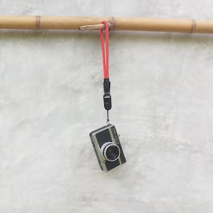 Puede incluir: Una cámara vintage en blanco y negro con una correa de muñeca roja cuelga de una caña de bambú contra una pared gris. La lente de la cámara está centrada y la correa está sujeta a la caña.