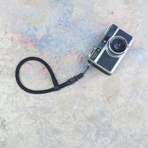 May include: A black and silver Fujifilm Compact camera with a black wrist strap. The camera has a lens and several buttons. The wrist strap is looped and attached to the camera. The camera is on a light gray surface.