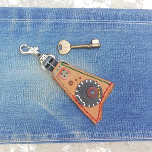 Op de afbeelding: Een handgemaakte leren sleutelhanger met een metalen sluiting, met een bruin leren ontwerp met kleurrijke stiksels en een knoopdetail. Een vintage messing sleutel ligt in de buurt. De items liggen op een blauwe denim achtergrond.