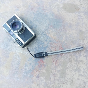 Pode incluir: Uma câmera de filme Fujifilm Compact 35mm vintage com corpo preto e prata e uma pulseira de pulso. A câmera tem uma lente e um flash. A pulseira é preta e branca.