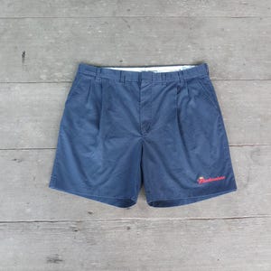 May include: Navy blue shorts with pleats and a button closure. The shorts have a small red "Budweiser" logo embroidered on the lower right leg. The shorts are laid flat on a weathered wooden surface.