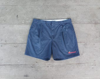 May include: Navy blue shorts with pleats and a button closure. The shorts have a small red "Budweiser" logo embroidered on the lower right leg. The shorts are laid flat on a weathered wooden surface.